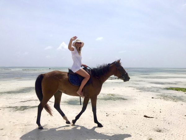 Tantra A woman riding a horse across the sandy beach, framed by the ocean breeze.