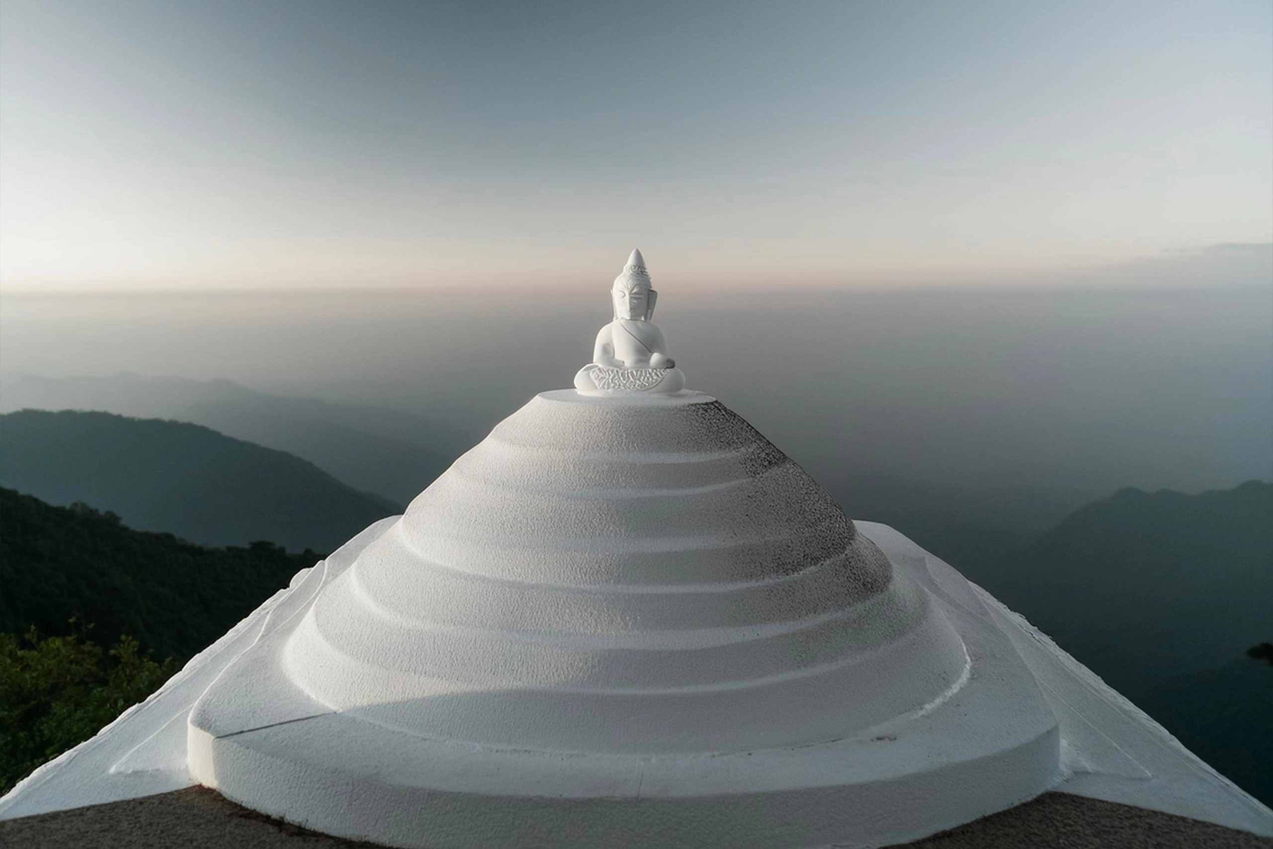 A white Buddha statue sits atop a layered stupa, overlooking a serene mountain landscape, symbolizing inner peace and spiritual ascension