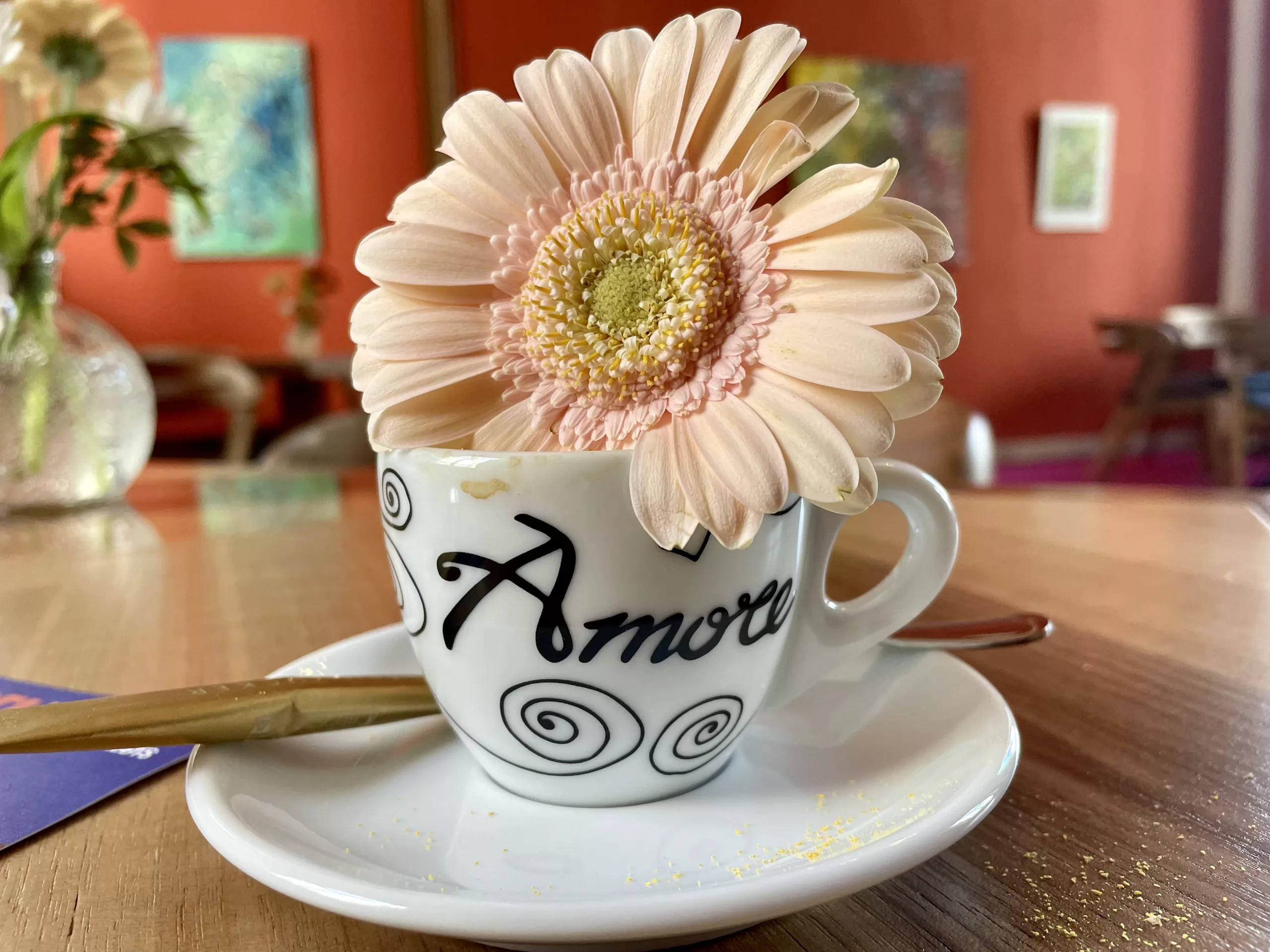 Tantra A cup of coffee with the words “With Love” written in the foam, next to a delicate pink daisy, symbolizing tenderness, awakening, and heartfelt presence
